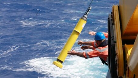 Crew member in an orange suit and blue helmet dropping a yellow Argo float from a ship into the sea