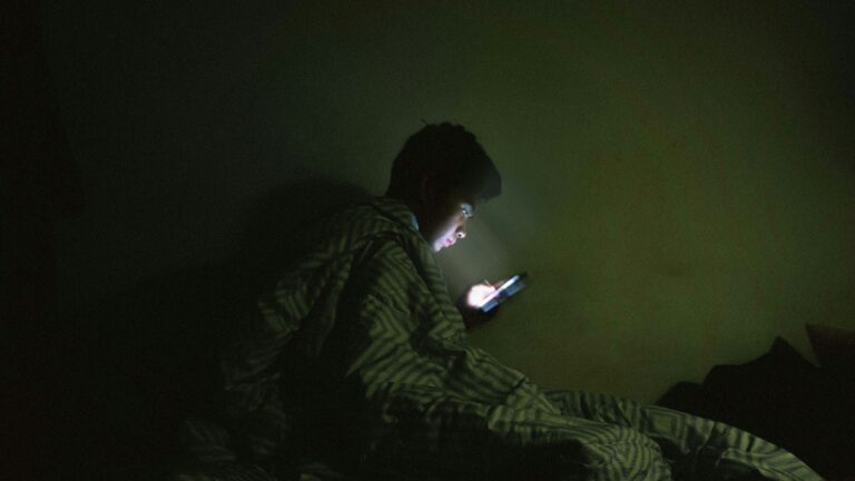 Person wrapped in a blanket in bed at night, looking at a smartphone