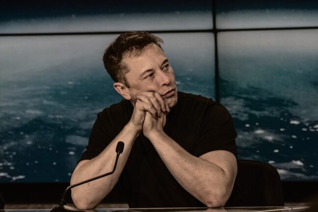Elon Musk sitting at a press conference table with his hands clasped under his chin