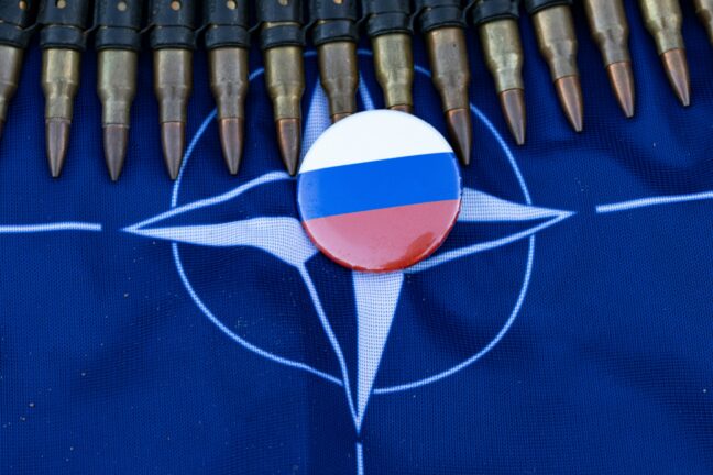 Badge with the Russian flag placed on a NATO flag, framed by a belt of rifle ammunition