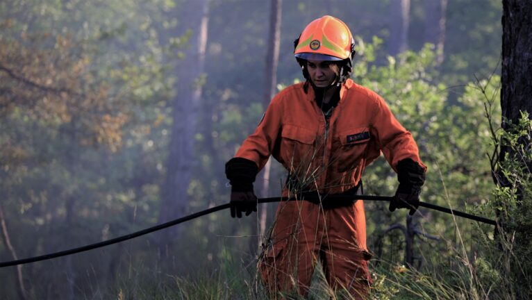 A firefighter in an orange uniform and helmet pulling a hose through a forest clearing