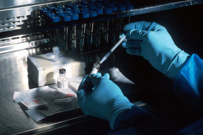 A researcher in blue gloves uses a syringe to draw liquid from a small vial in a laboratory setting