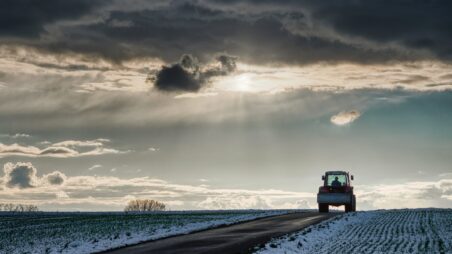 A tractor driving along a snow-covered farm road under a dramatic, cloudy winter sky