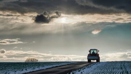 A tractor drives along a snow-lined road under dark storm clouds with sunlight breaking through