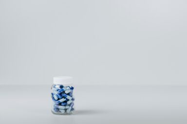 A small glass jar filled with blue and white capsules on a plain grey background