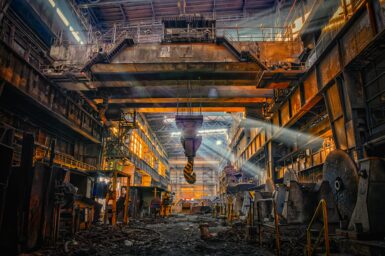 The rusted interior of a disused steel factory hall, with an overhead crane hook hanging in the centre