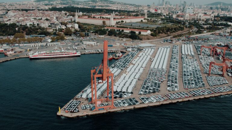 Aerial view of rows of new cars parked at a shipping port, with a crane and city skyline in the background