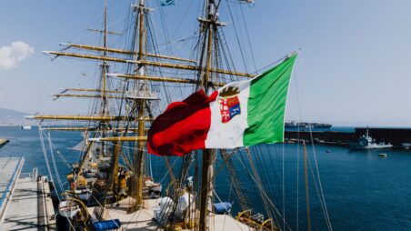Italian naval ensign flying aboard a training ship, with a container vessel in the background
