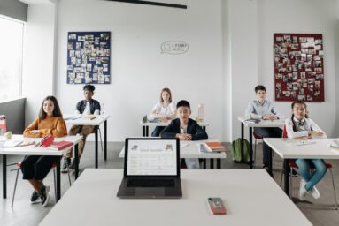 Group of school pupils sitting at desks in a bright classroom, with a laptop showing a school timetable in the foreground