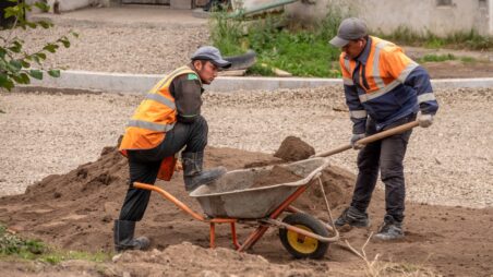 Two workers at a construction site, with one shovelling sand into a wheelbarrow