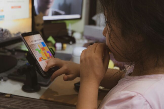 A young girl uses a tablet at a desk while biting her fingernail.