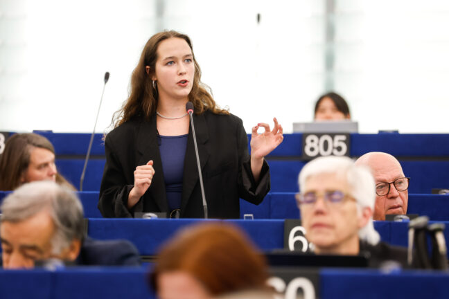 Lena Schilling stands and gestures as she speaks into a microphone during a plenary session of the European Parliament, with lawmakers seated around her