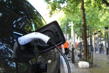 A dark car charging from a public charging cable on a tree-lined street