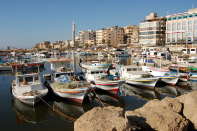 Fishing boats moored in the harbour of Tartus, with the city’s waterfront and a minaret behind them