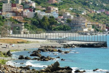 Border fence running along a beach and into the sea, with houses on a green hillside behind it