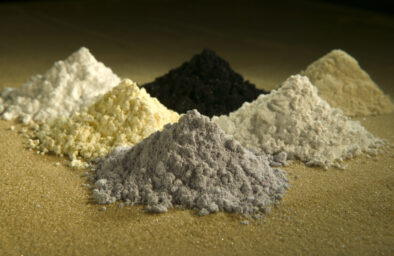Six samples of rare earth oxide powders in small dishes, including praseodymium, cerium, lanthanum, neodymium, samarium, and gadolinium