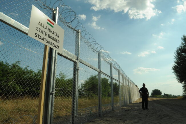 Razor-wire fence along Hungary’s southern border with a trilingual “State border” sign and a guard in silhouette