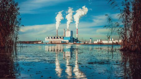Industrial facility with tall chimneys releasing plumes of smoke, reflected in the still water of a lake