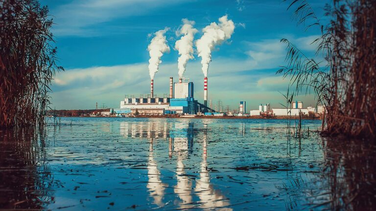 Industrial facility with tall chimneys releasing plumes of smoke, reflected in the still water of a lake