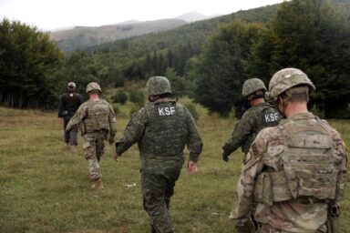 Kosovo Security Force members and US soldiers walking across a grassy field towards forested hills