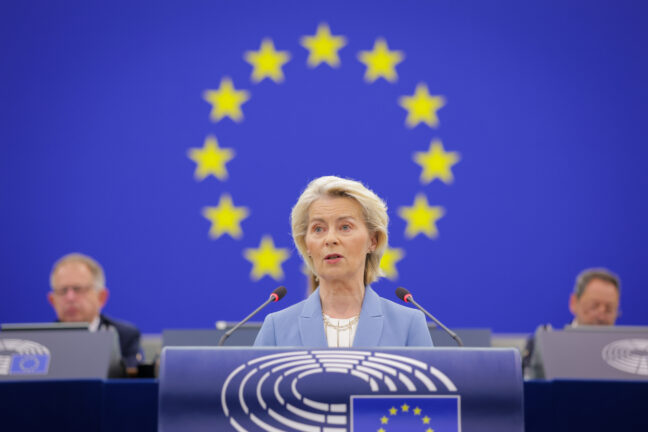 Ursula von der Leyen speaking at the podium of the European Parliament in Strasbourg, with the EU flag behind her