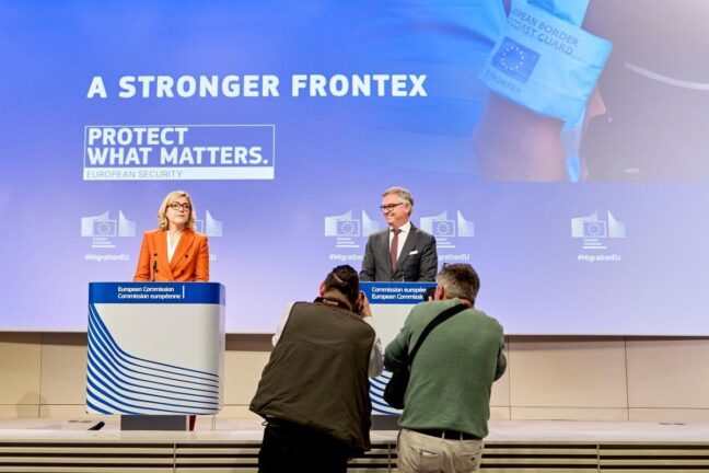 Two European Commission officials speak at podiums in front of a banner reading “A stronger Frontex. Protect what matters”, with photographers in the foreground