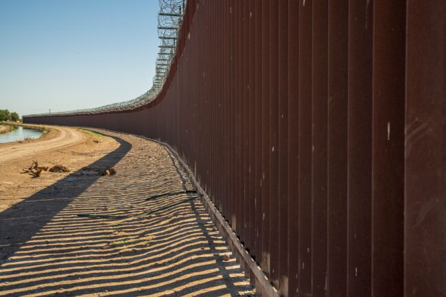 Tall rust-coloured steel border wall topped with razor wire, casting striped shadows on sandy ground