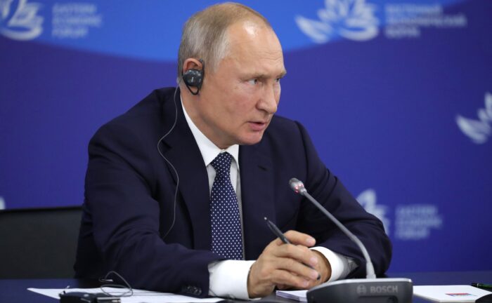Russian President Vladimir Putin with an interpreter earpiece speaking at a table with a microphone, in front of a blue Eastern Economic Forum backdrop