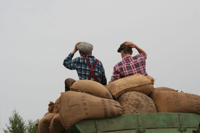Two men in checked shirts sit on top of sacks loaded on a cart, shading their eyes as they look into the distance