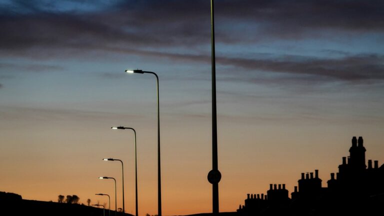 A row of lit streetlamps against a dawn sky, with the silhouettes of rooftops and chimneys